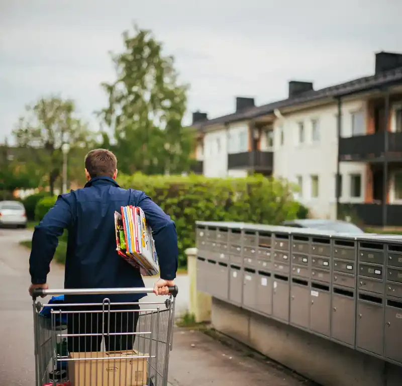 Postijakaja sinisessä takissa työntää jakelukärryä suomalaisella asuinkadulla postilaatikoiden ohi, käsivarren alla mainoslehtisiä.